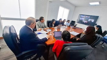 Un grupo de personas se reúne alrededor de una mesa de conferencia con computadoras portátiles frente a una pantalla con videollamada
