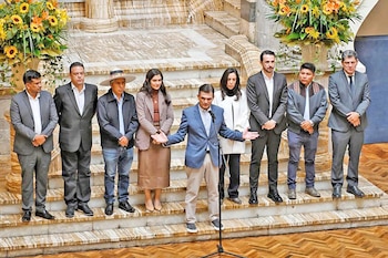 Diez personas en un recinto formal, un hombre habla por un micrófono, ocho hombres y dos mujeres lo escuchan de pie en una escalera, con arreglos florales