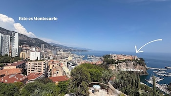 Vista panorámica de una ciudad costera con edificios, montañas, un puerto con yates y el mar. Se aprecian textos "Esto es Montecarlo" y "Esto es Mónaco"