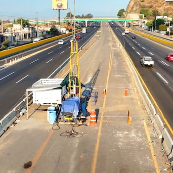 Vista aérea de equipos de construcción amarillos y azules en el carril central de una autopista, con tráfico de vehículos y un puente verde al fondo