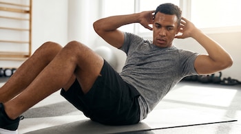 Un hombre sudoroso de tez oscura haciendo abdominales sobre una colchoneta gris en un gimnasio iluminado por el sol, con las manos detrás de la cabeza.