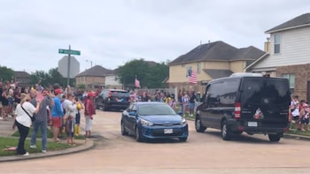 Calle residencial llena de gente, ondeando banderas de EE. UU. en las aceras. Una van negra y un coche azul circulan. Casas y árboles en el fondo