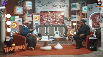 Un estudio futurista con dos hombres en sillones, mesas con trofeos y objetos. Pantallas retro, luces circulares y una camiseta de fútbol visible