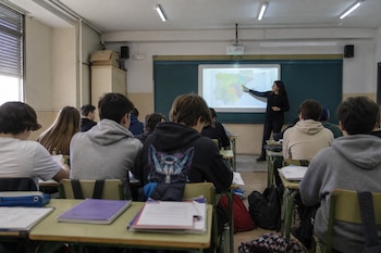 Alumnos de espaldas sentados en sus pupitres miran a una profesora que señala un mapa proyectado al fondo del aula.
