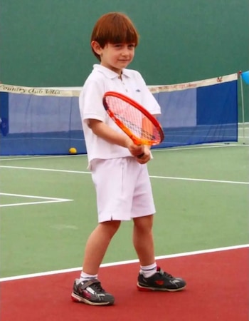 Niño con cabello castaño claro, vestido con polo y shorts blancos, sostiene una raqueta de tenis naranja en una cancha. Se ve una red al fondo