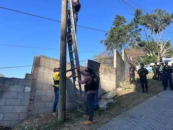 Un hombre en escalera sobre poste de madera, otros tres lo asisten. Al fondo un muro de piedra, cables eléctricos aéreos y un cielo azul claro visible