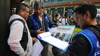 Tres hombres conversan en una calle concurrida; uno, vestido con chaleco azul, muestra un folleto sobre el correcto uso de la bandera nacional