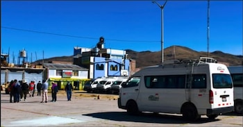 Varias furgonetas de transporte blancas están estacionadas en un patio abierto, con personas reunidas cerca y edificios coloridos al fondo bajo un cielo azul claro