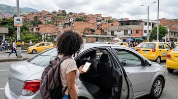 Persona con mochila y cabello rizado subiendo a un coche plateado, con el móvil mostrando la app DiDi. Un conductor dentro. Casas en ladera y taxis amarillos.