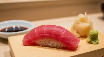 Primer plano de un akami nigiri sushi con atún rojo sobre una tabla de madera clara, junto a un tazón de salsa de soja, jengibre y wasabi.