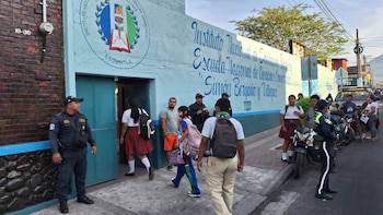 Oficiales de seguridad uniformados están de pie frente a un edificio escolar azul con estudiantes entrando y saliendo, junto a una calle con vehículos