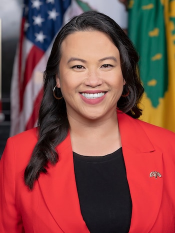 Sheng Thao sonríe con una chaqueta roja y un broche en la solapa, ante la bandera de Estados Unidos y una bandera verde y amarilla