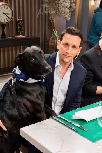 Hombre de traje y longdog negro con pañuelo azul sentados en una mesa. Hay cubiertos, un vaso, un reloj y objetos decorativos