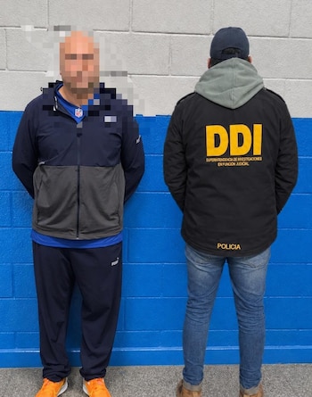 Un hombre con el rostro pixelado se encuentra de pie junto a un policía de espaldas con una chaqueta que tiene las siglas DDI