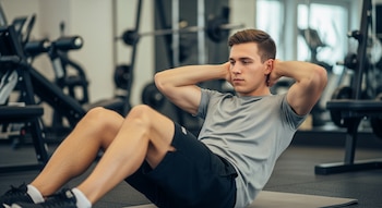 Jovem de pele clara fazendo abdominais em um tapete cinza na academia. Ele veste uma camiseta cinza e shorts pretos.