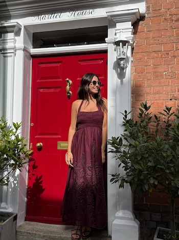 Pampita de pie, sonriendo y mirando hacia la derecha, frente a una puerta roja con molduras blancas y detalles dorados. Viste un vestido largo bordado y gafas de sol