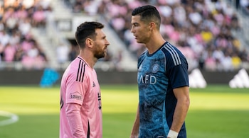 Lionel Messi con camiseta rosa y Cristiano Ronaldo con camiseta azul se miran fijamente en un campo de fútbol con público al fondo. Ambos tienen barba y pelo oscuro.