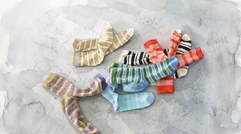 Calcetines con patrones de rayas en colores amarillo, morado, verde, rojo y negro sobre un fondo gris con textura de acuarela.