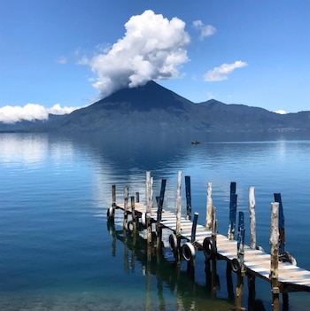 Un muelle de madera en primer plano se extiende sobre un lago azul claro. Al fondo, un volcán grande con una nube blanca encima se alza bajo un cielo azul
