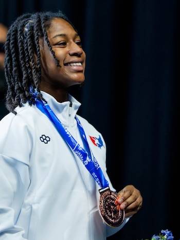 Una joven con trenzas viste una chaqueta blanca deportiva y sostiene una medalla colgada de una cinta azul