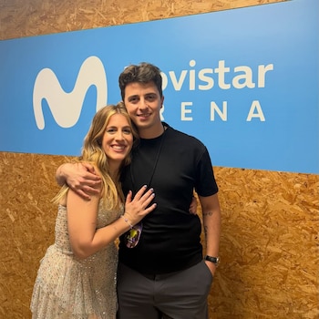 Hombre y mujer sonrientes posan abrazados, la mujer con vestido brillante y el hombre con camiseta negra, frente a un letrero de Movistar Arena