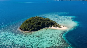 Vista aérea de una isla tropical densamente arbolada con una playa de arena blanca. El agua circundante es de un turquesa claro, revelando arrecifes de coral, y se convierte en un azul profundo más lejos.