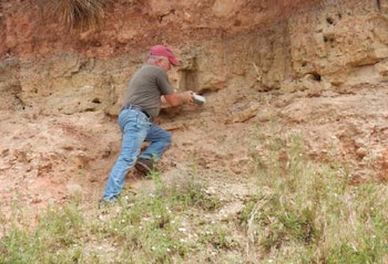 Un hombre con gorra roja y pantalones azules sostiene un objeto frente a una pared de tierra arcillosa