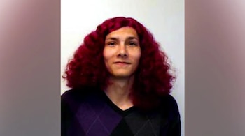 Retrato de hombre con cabello largo rizado de color rojo, con suéter oscuro de cuello en V y patrón de rombos