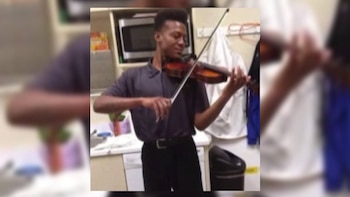 Joven con camisa polo gris oscuro tocando un violín marrón. Viste pantalones oscuros. Al fondo se ven estantes blancos y un microondas plateado