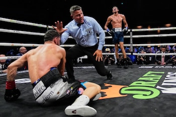 Un boxeador en la lona, un árbitro de pie con la mano extendida y otro boxeador de pie al fondo dentro de un cuadrilátero de boxeo