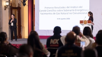 Dos mujeres en un escenario. Una con micrófono, la otra en un podio frente a una pantalla con texto. Público sentado en primer plano borroso.