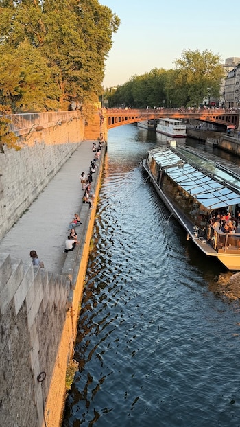Un barco turístico grande navega en un río urbano. Varias personas sentadas en una orilla de piedra. Un puente de metal y árboles en el fondo