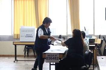 Un miembro de mesa de la ONPE entrega documentos a otro miembro sentado en una mesa con el rótulo "MESA N° 036482" y una cabina de votación