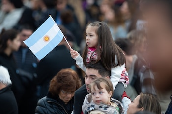 Una niña con cabello largo y oscuro es cargada sobre los hombros de un adulto, sosteniendo una pequeña bandera argentina. Otros adultos la rodean en el fondo