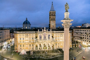 Vista aérea nocturna de la Basílica Papal de Santa María la Mayor en Roma, con su fachada iluminada y el campanario, junto a la columna de la Salus Populi Romani y edificios circundantes