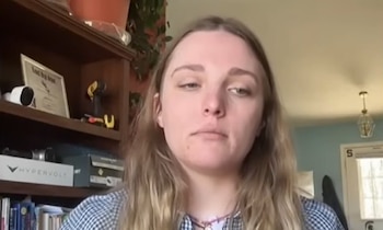 Primer plano de Rachel Gil, una mujer de cabello rubio, mirando directamente a la cámara con una camisa a cuadros. Al fondo, una estantería de madera