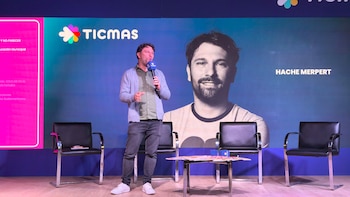 Hache Mempert de pie en un escenario, sosteniendo un micrófono, con una pantalla grande detrás que muestra el logo de Ticmas y su retrato