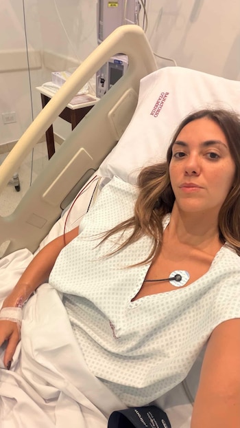 Floppy Tesouro recostada en una cama de hospital, vistiendo una bata médica, con una vía intravenosa en el brazo izquierdo y un electrodo en el pecho