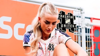 Mujer sentada en césped artificial con camiseta deportiva y medias negras, ajusta su calzado. Fondo con muro naranja y valla. Logotipos Hashtag United y Kelci-Rose
