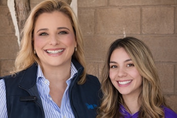 Primer plano de dos mujeres sonrientes: una rubia con chaleco azul y camisa a rayas, y otra morena con cabello largo y camisa morada, frente a una pared de ladrillos.
