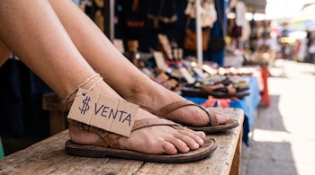 Primer plano de dos pies de mujer con sandalias de cuero y una etiqueta de cartón en el tobillo que lee "VENTA" con un símbolo de dólar. Fondo de mercado.