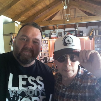 Primer plano de dos hombres; uno con barba y camiseta negra, otro con gafas de sol, gorra blanca y negra con un logo, y camisa a cuadros