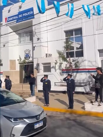 Edificio de la Central de Policía con banderas argentinas. Varias personas, incluidas oficiales de policía, saludan al paso de un vehículo plateado