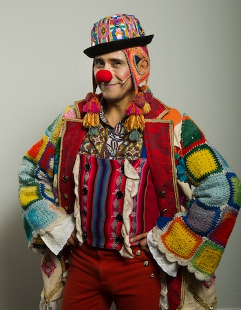 Hombre con nariz de payaso, maquillaje en mejillas, sombrero colorido con detalles andinos, chaqueta multicolor con parches de ganchillo, chaleco a rayas y pantalón rojo