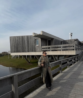 Una mujer de abrigo largo de piel, pantalón oscuro y gafas de sol en pasarela de madera. Detrás, un edificio de madera elevado y un cuerpo de agua