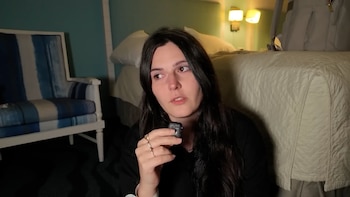 Una mujer de cabello oscuro con blusa negra sostiene un dispositivo electrónico en su mano derecha. La rodean un sillón a rayas azules y blancas, una cama y una mochila
