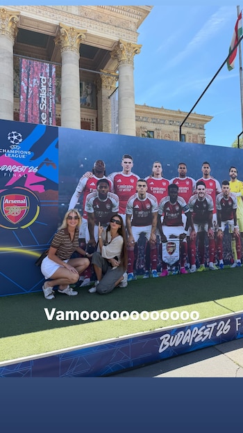 Dos mujeres posan sonrientes frente a un cartel del Arsenal y la Champions League en Budapest. Un edificio clásico y la bandera de Hungría al fondo