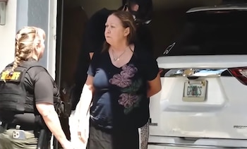 Mujer con blusa oscura y esposas es escoltada por una oficial de sheriff con chaleco negro frente a un vehículo blanco con placa de Florida