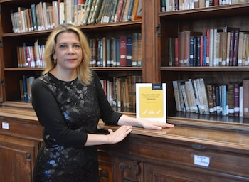 Mujer con cabello rubio, jersey negro con encaje, pendientes dorados, sentada ante estanterías de madera llenas de libros, con un libro amarillo
