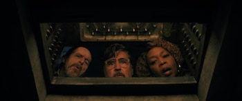 Tres personas se asoman desde una abertura oscura. Un hombre con gafas y barba en el centro, flanqueado por un hombre barbudo y una mujer de cabello rizado